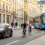 Les 8 équipements pour déplacements urbains qui simplifient le quotidien Rue urbaine animée le matin avec un vélo, un homme et une femme à pied, un autobus de transport public et une voiture compacte.