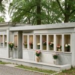 Columbarium contemporain en pierre dans un cimetière arboré de Belgique, orné de petits vases fleuris sous une lumière douce.